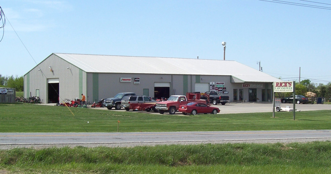 Exterior view of a large gray warehouse, possibly serving as a rental hub, with multiple garage doors and signs. Several cars and equipment are parked in front. A sign on the lawn reads 