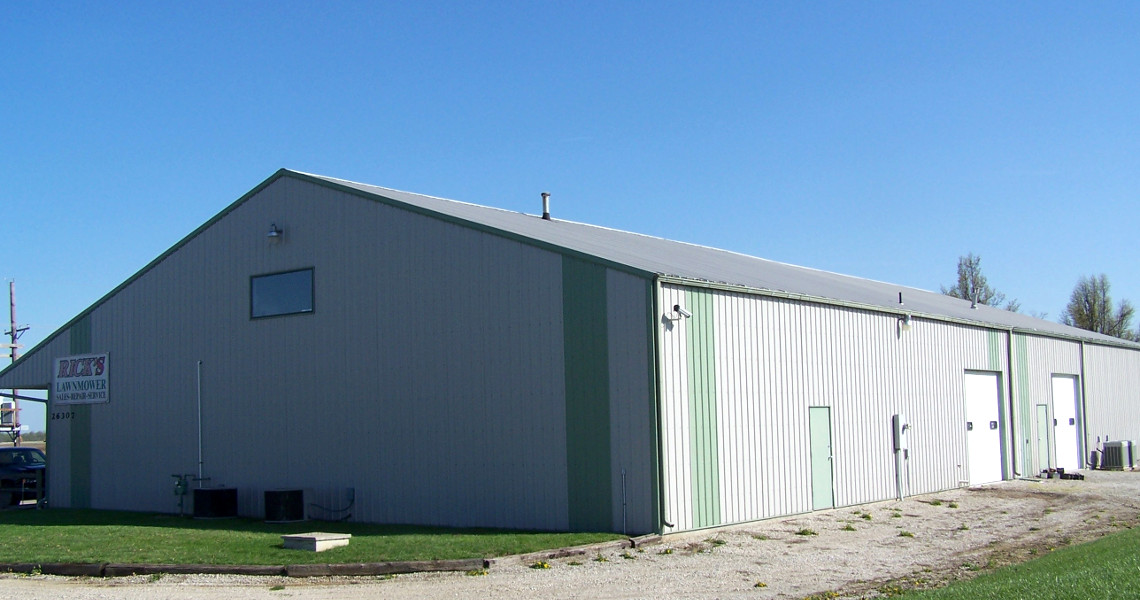 A spacious, light gray warehouse with green trim and a peaked metal roof sits on a grassy area with a gravel driveway, perfect for rental use. It's a clear day with a blue sky in the background.