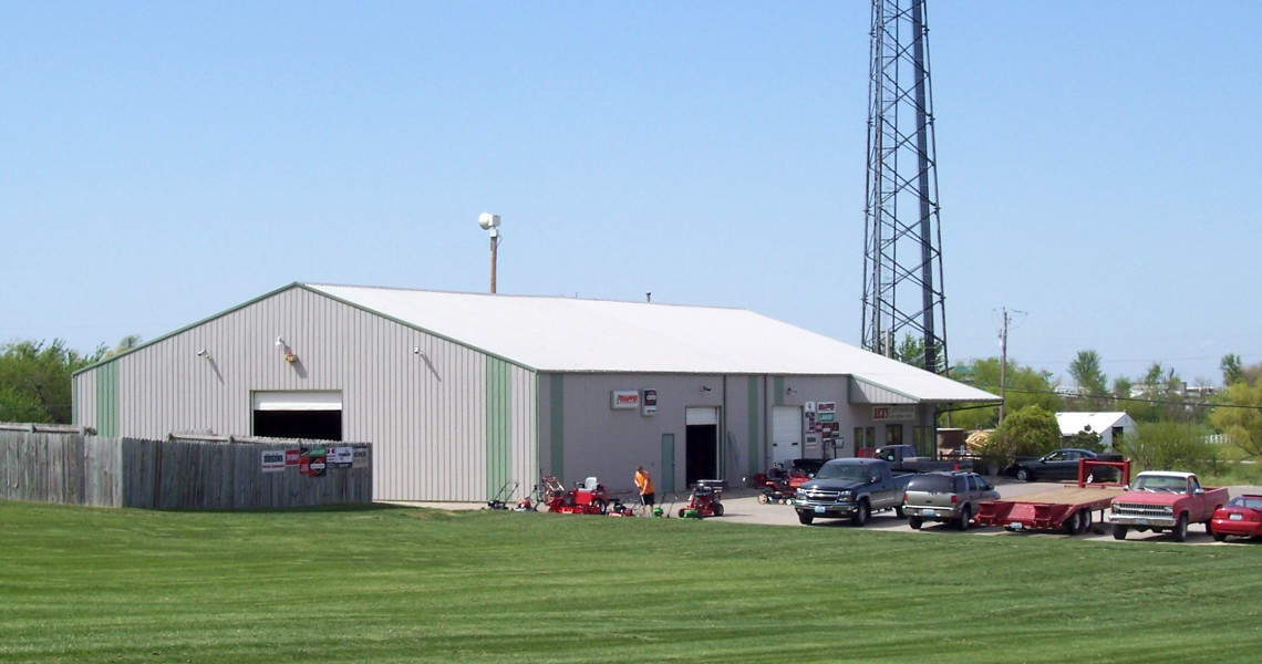 A metal warehouse building with a white roof, located next to a tall radio tower, serves as both a rental hub and storage space. Several vehicles and outdoor equipment are parked in front, while the surrounding area features well-maintained grass and trees in the background.