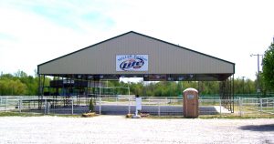A large open pavilion with a metal roof labeled "Miller Pavilion Lite" stands in a gravel area, reminiscent of a fair building. A portable toilet is on the right side, and county fair-style picnic tables are underneath. Trees and greenery are visible in the background.