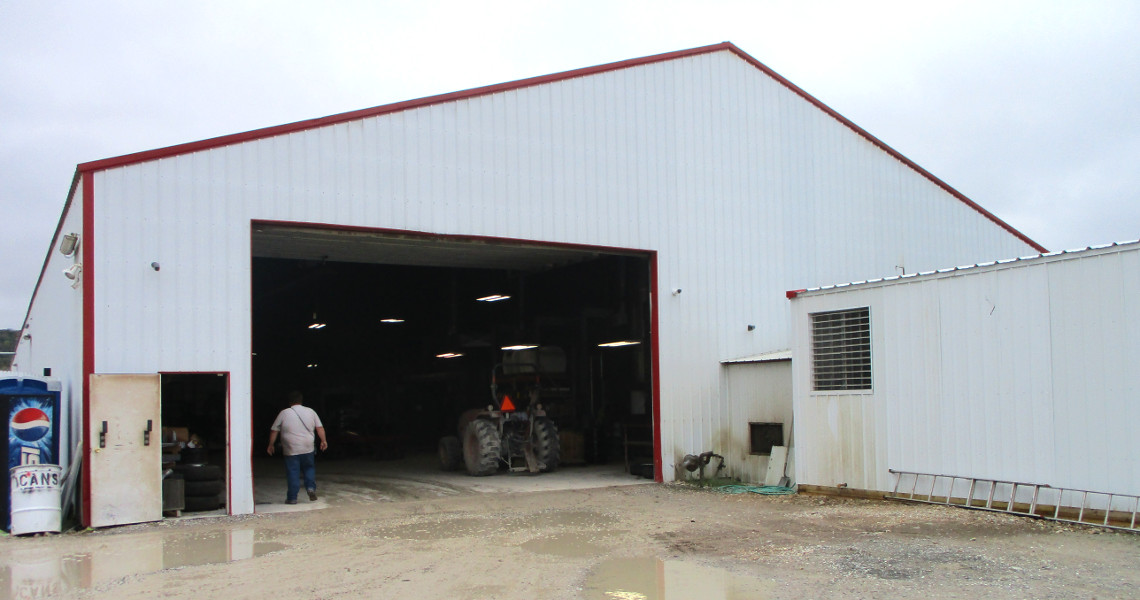 A large white barn with a red-trimmed roof and an open garage door reveals a tractor and a few trailers inside. A person stands near the entrance, where the ground is muddy with a few puddles. A ladder leans against the building, and a Pepsi vending machine is visible.