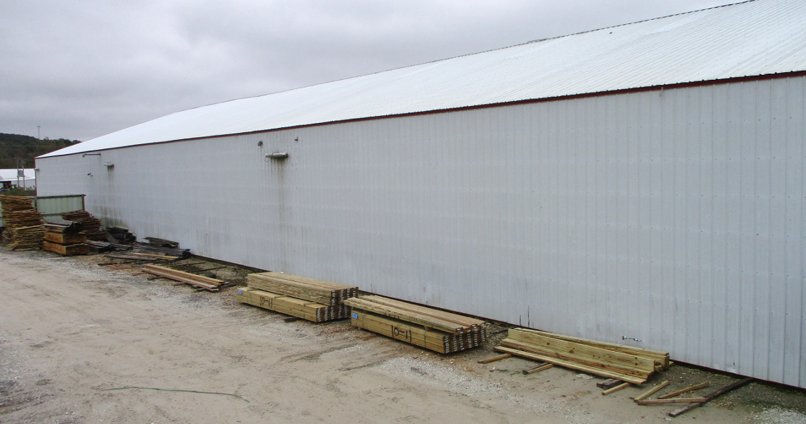 A large, white metal warehouse with a sloped roof is seen on a cloudy day. Trailers are parked nearby, and stacks of wooden planks are neatly arranged along the building's exterior on a gravel surface. A dirt path runs beside the warehouse.