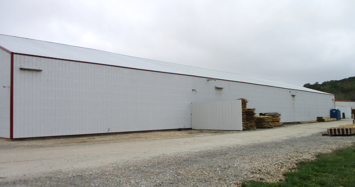 A large, long white industrial warehouse with a sloped roof stands against a cloudy sky. There's a gravel road in front and stacks of trailers and wooden pallets along the side of the building, hinting at TrailerMan's bustling operations.