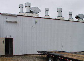 A long white industrial building, with multiple ventilation pipes on the roof, highlights the scene. An open door is visible on the left side. In the foreground, a TrailerMan flatbed trailer is parked on a gravel surface under an overcast sky.
