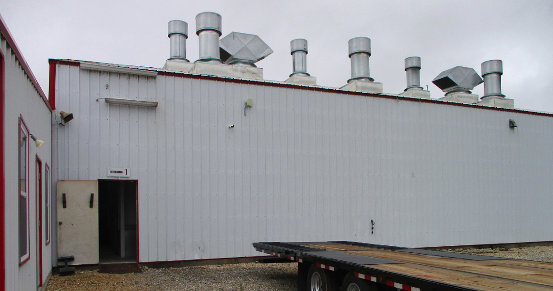 A long white industrial building, with multiple ventilation pipes on the roof, highlights the scene. An open door is visible on the left side. In the foreground, a TrailerMan flatbed trailer is parked on a gravel surface under an overcast sky.
