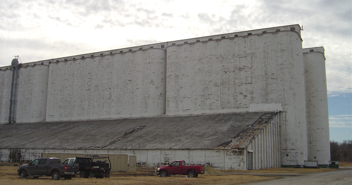 A large, weathered white concrete building stands tall, featuring a grain elevator with multiple cylindrical silos and a long sloped roof. Several vehicles are parked in front, enhancing the bustling scene on a partly cloudy day.