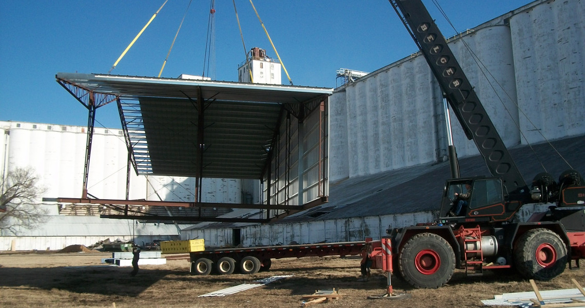 A large crane lifts a metal roof section at a construction site, surrounded by tall industrial silos and an imposing building. The structure is being carefully placed onto a flatbed truck. The scene unfolds on a clear day under a blue sky.