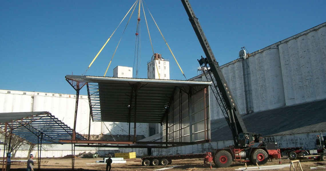 A crane lifting a large metal structure at a construction site, with workers observing the scene. In the backdrop, tall white industrial buildings rise under a clear blue sky, resembling an intricate urban elevator to the heavens.