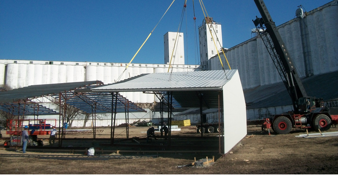 A crane lifts a large metal roof panel to be installed on a building frame at a bustling construction site. Several workers are guiding the panel into place on a clear day, with towering buildings and large industrial structures visible in the background.