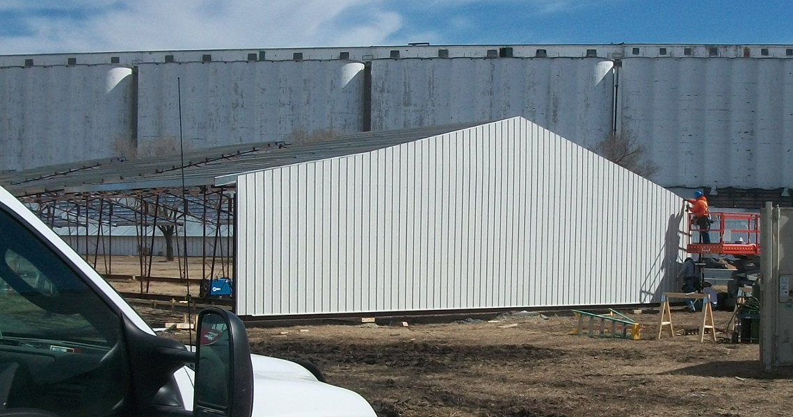 A construction site buzzes with activity as an orange-shirted worker operates a lift, expertly attaching panels to the white metal-paneled building. Nearby, vehicles and materials are scattered across the foreground, while a towering industrial structure looms in the background.
