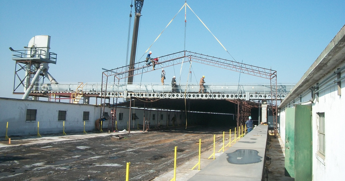 Workers are using a crane to construct a large metal framework over an industrial building. The structure, resembling an open-roofed elevator shaft, hosts several workers on top. Yellow safety posts line the pathway below. The sky is clear and bright.