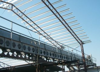 A steel framework of a building under construction, featuring metal beams and girders against a clear blue sky. The structure is partially completed, with open areas revealing scaffolding, support beams below, and space for an elevator.