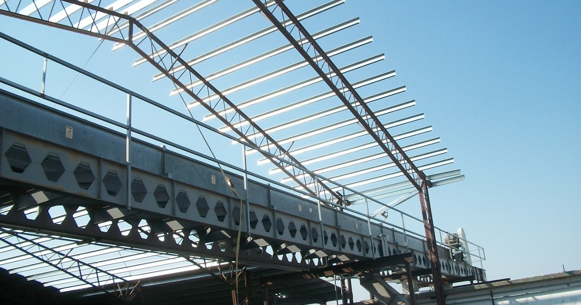 A steel framework of a building under construction, featuring metal beams and girders against a clear blue sky. The structure is partially completed, with open areas revealing scaffolding, support beams below, and space for an elevator.