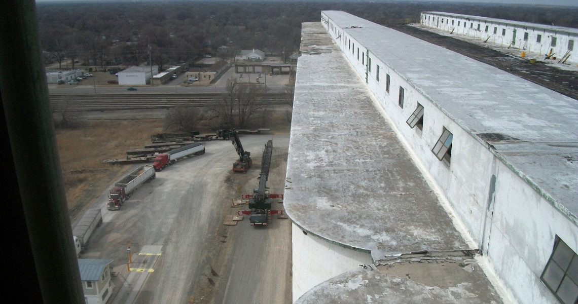 A view from a high building reveals an industrial area with parked trucks and trailers. Amidst the scene is a towering, weathered concrete structure resembling an old elevator, its windows surveying adjacent roads and the distant landscape beneath a cloudy sky.
