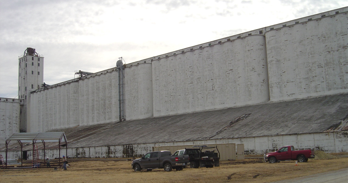 A large white grain elevator building with multiple storage silos is surrounded by parked vehicles on a dry grass field. The sky is overcast, and there are some small structures and utility buildings nearby.