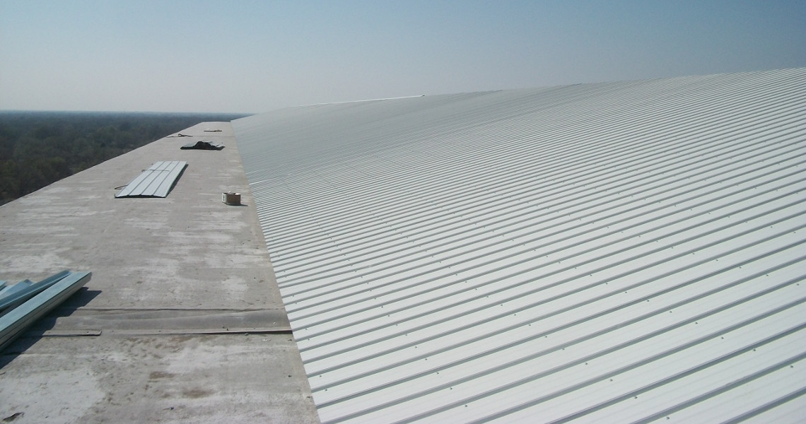 A large white corrugated metal roof stretches into the distance under a clear blue sky, atop a bustling building. Several metal sheets and tools are scattered across the roof, hinting at ongoing construction or maintenance work. The horizon is faintly visible.