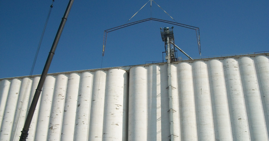 A series of tall, white grain silos stand under a clear blue sky, resembling a building skyline. A crane is lifting a metal structure above the silos while an elevator platform carries a worker overseeing the operation.