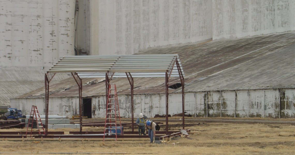 Two workers assemble a metal frame for a small structure on dry grass, with an old building looming in the background. A ladder and construction materials are nearby, suggesting future expansion possibly involving an elevator installation.