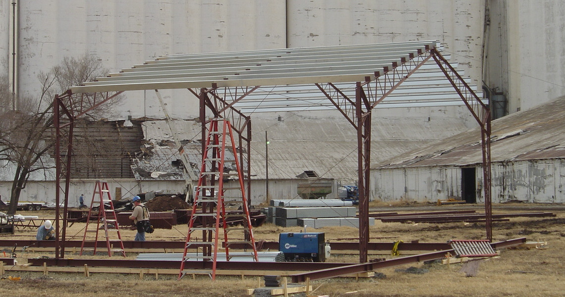 A metal structure under construction stands in an open field, resembling a nascent building. It features a framework of red beams, partially completed roofing, and an elevator shaft forming. Two ladders are positioned nearby, and construction materials are scattered around. In the background, industrial buildings are visible.