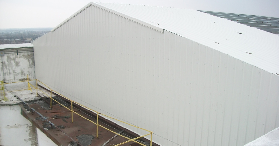 A large, white industrial building under construction with a slanted roof features prominently against an overcast sky. Yellow scaffoldings surround the structure, hinting at future elevator installations. The setting suggests it's located in a bustling industrial area.
