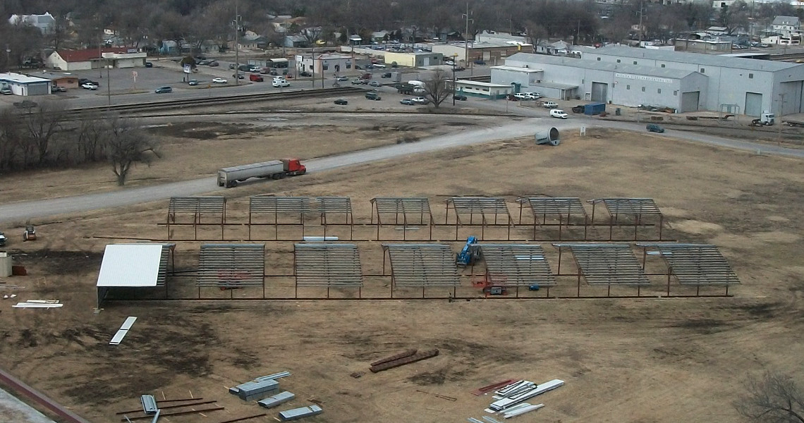 An aerial view of a construction site features multiple metal frame structures being assembled on a large plot of land. Several piles of materials are scattered around. A crane, truck, and elevator are visible, with buildings and roads in the background.
