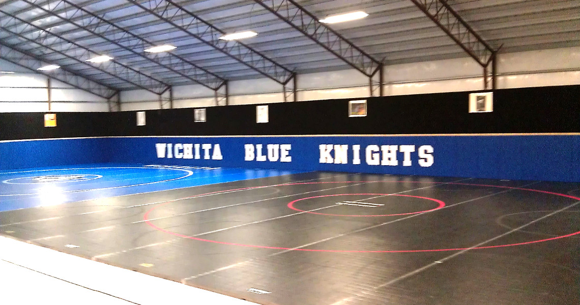An indoor wrestling gym with a blue and black mat featuring red and white circles. The wall proudly displays 