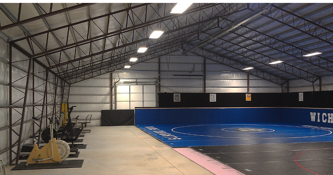 The Blue Knight Facility boasts a large indoor gymnasium with wrestling mats spread across the floor. Its high metal ceiling and walls encompass exercise equipment, like an air bike, neatly lined along the side. Bright lights illuminate every corner of this impressive facility.
