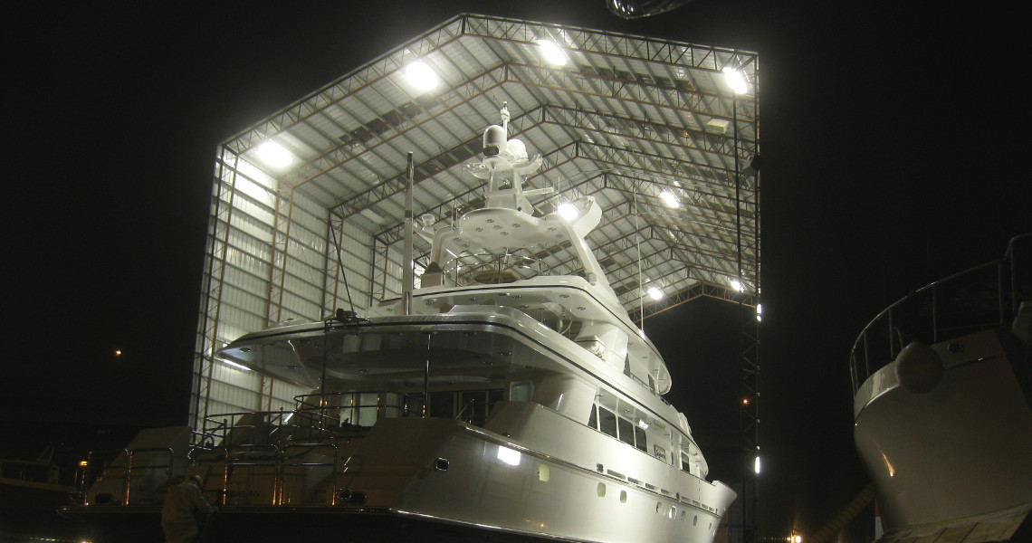 A large, white yacht is docked inside an illuminated, open-roof shipyard at night. The maritime structure shelters the vessel, with lights highlighting its features. Other boats are partially visible on the sides, adding to the nautical atmosphere.