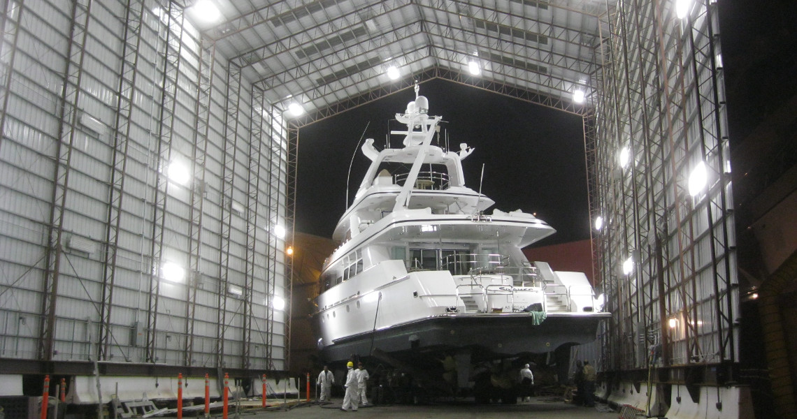 A large white yacht sits proudly inside The Shipyard hangar at night, bathed in light as several workers bustle around its base. The hangar's open ends frame a clear night sky, adding to the majestic scene.