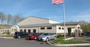 A commercial building serves as Calo's headquarters, with a sign proudly displaying "Calo." Two cars are parked in front, an American flag waves on a pole, and the structure is nestled against a backdrop of trees beneath a clear blue sky.