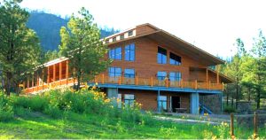 This classic retreat features a large wooden building with multiple windows and a wraparound balcony, surrounded by lush greenery and pine trees. Sunflowers bloom in the foreground, while a majestic mountain rises in the background under a clear blue sky.