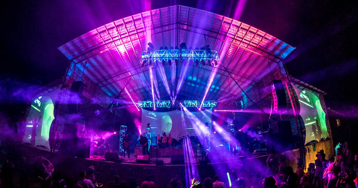 A vibrant nighttime concert scene transforms into a dynamic disco environment with colorful lights illuminating the large outdoor stage. Performers shimmer under beams of pink, purple, and blue as the energized crowd watches from below, enhancing the lively atmosphere.