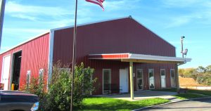 A large red barn-style building with a white roof and multiple windows sits under a clear blue sky. An American flag waves in the foreground, while a Caskey Trucking vehicle is partially visible on the left, surrounded by lush green grass.