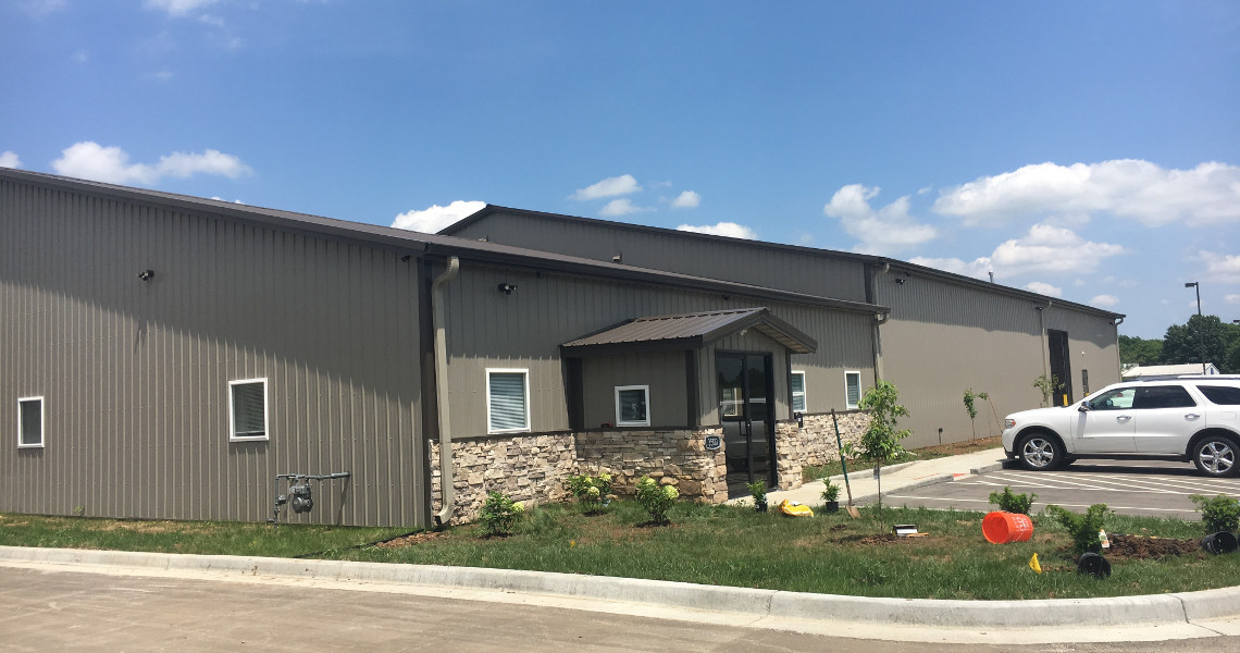 A modern industrial building featuring a stone facade and metal siding stands under a clear blue sky. The entrance, with its small overhang, invites visitors as a white SUV is parked nearby. Newly planted landscaping nods to recent construction efforts, enhancing the brokers' appeal of this space.