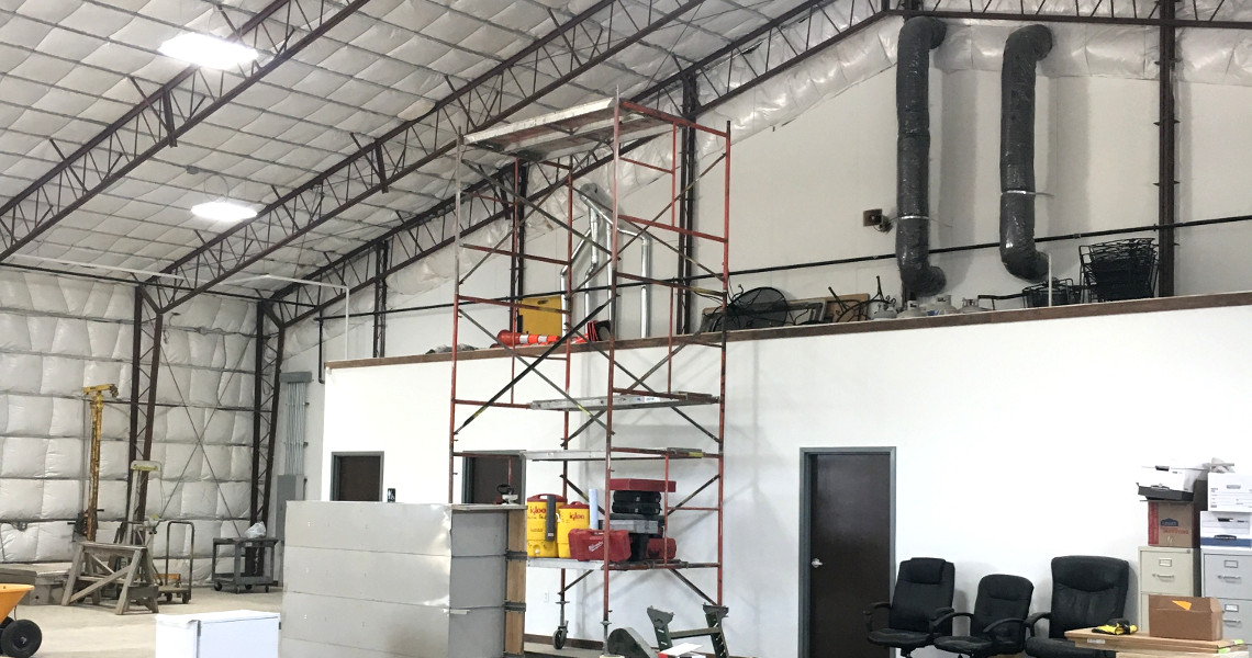 A large industrial warehouse with a high ceiling, filled with construction scaffolding, office chairs, filing cabinets, and various tools and equipment. The walls are insulated, and there are large ventilation pipes visible.