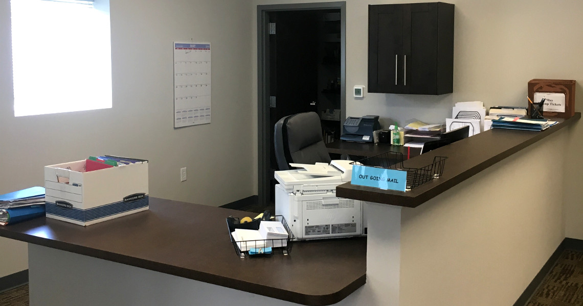 A tidy office reception area with a countertop desk containing a printer, organized papers, and office supplies. Behind the desk, a chair awaits brokers while cabinets and a wall calendar rest in the background. Natural light floods the space from a window.