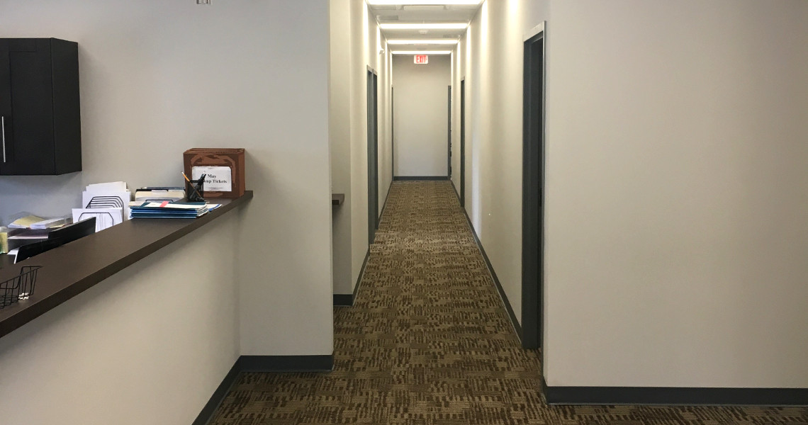 A well-lit office hallway with carpeted flooring. On the left, a reception counter featuring papers and a 