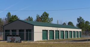 A row of John's storage units with green doors and a green roof is surrounded by a chain-link fence. A sign in front details rental sizes and rates. Tall trees stand in the background under a clear blue sky.