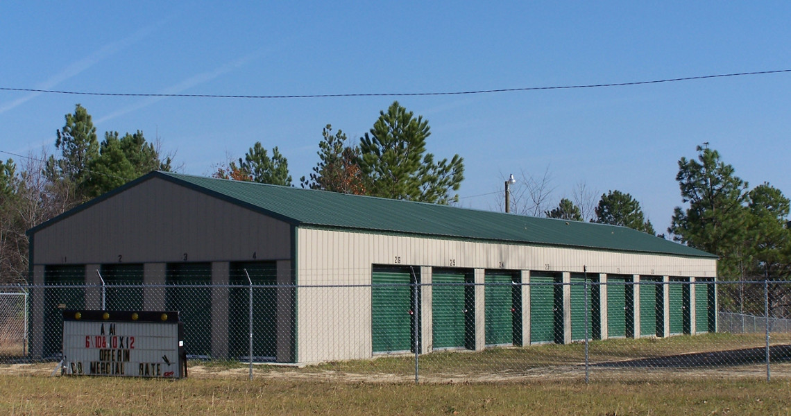 A row of John's storage units with green doors and a green roof is surrounded by a chain-link fence. A sign in front details rental sizes and rates. Tall trees stand in the background under a clear blue sky.