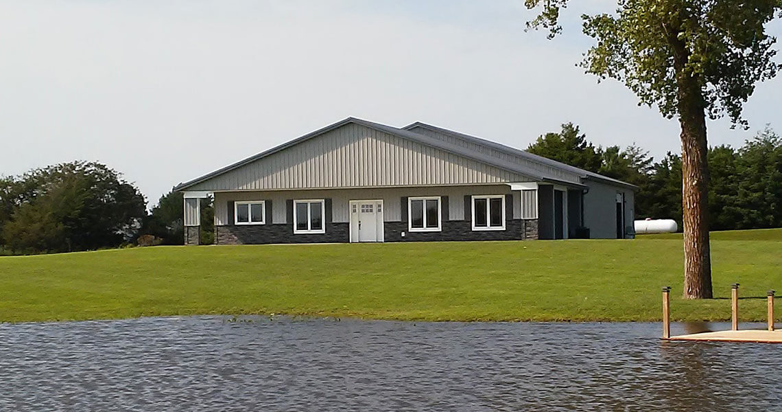 A large metal building with a gray roof and white doors sits on a grassy hill. There's a tree to the right and a body of water in the foreground. The sky is clear, and trees are visible in the background.