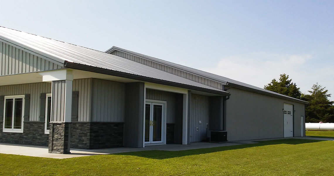 A modern gray building with a metal roof and stone accents. It features double glass doors, a large garage door, and is surrounded by a green lawn under a clear blue sky.