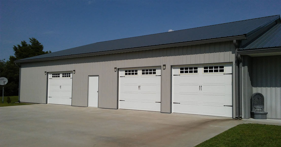 A large, gray metal building with a dark roof features three white garage doors and a single white pedestrian door. The area in front is a flat concrete driveway, bordered by grass and a small satellite dish to the left.