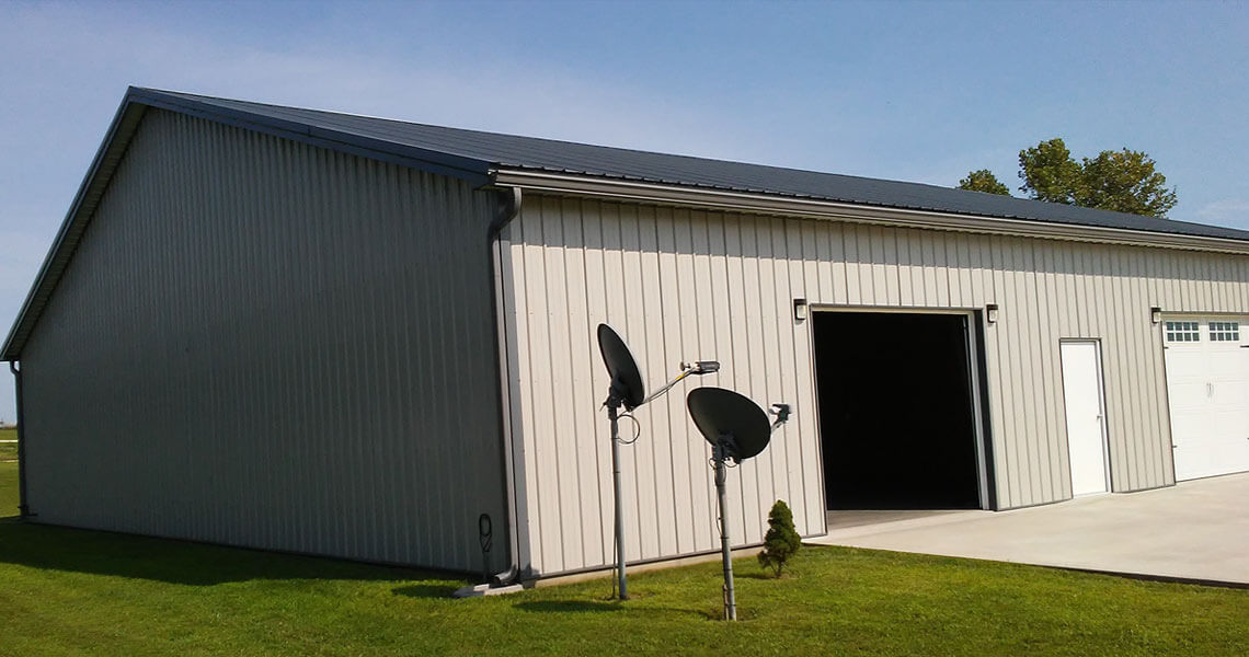 A large gray metal building with a partially open garage door and an adjacent closed door. Two satellite dishes are mounted on a pole in the grassy yard, and a small conifer tree stands nearby. The sky is clear and blue.