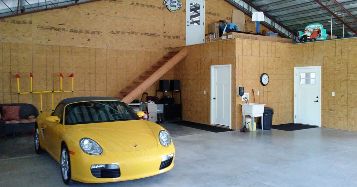 A spacious garage with a yellow sports car parked on the left. The interior features wooden walls, a staircase leading to an upper level, and a section with a white sink and two closed doors. A wall clock is visible above the sink.