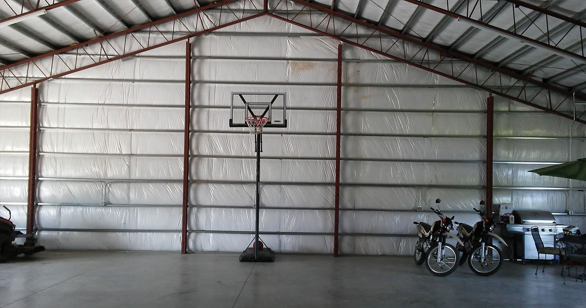 A large metal garage with a portable basketball hoop in the center. On the right, there are two motorcycles and a barbecue grill. The floor is concrete, and the walls and ceiling are made of sheet metal.