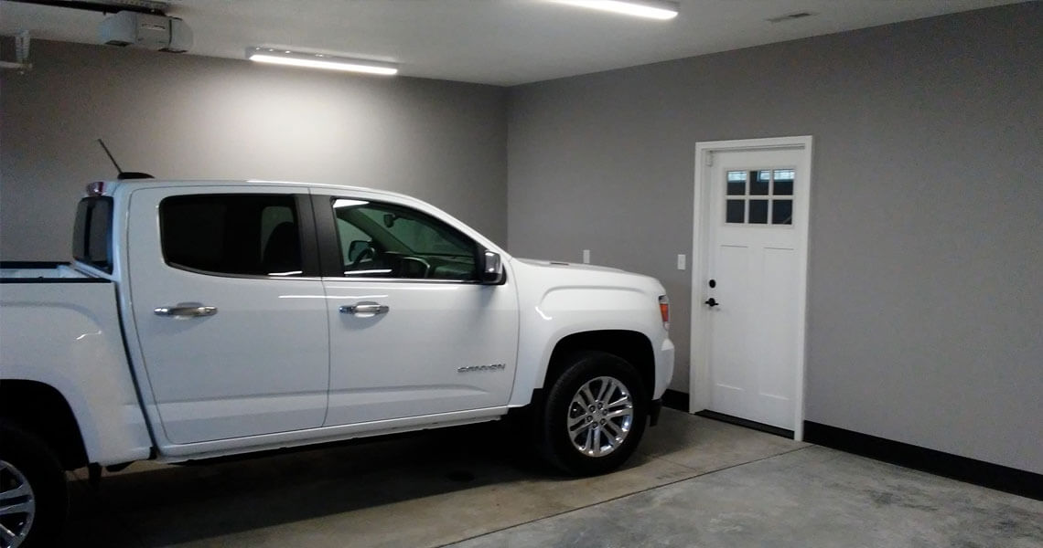 A white pickup truck is parked inside a clean, empty garage with gray walls. A white door with glass panels is visible on the right wall. The garage is well-lit with overhead lights.