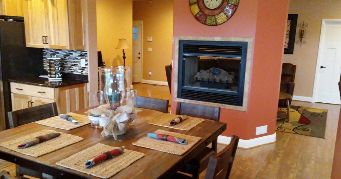 A cozy dining room features a wooden table set for six with woven placemats and cloth napkins. The room includes a decorative fireplace, warm-toned walls, modern kitchen cabinetry, and a wall clock. A separate sitting area is visible in the background.