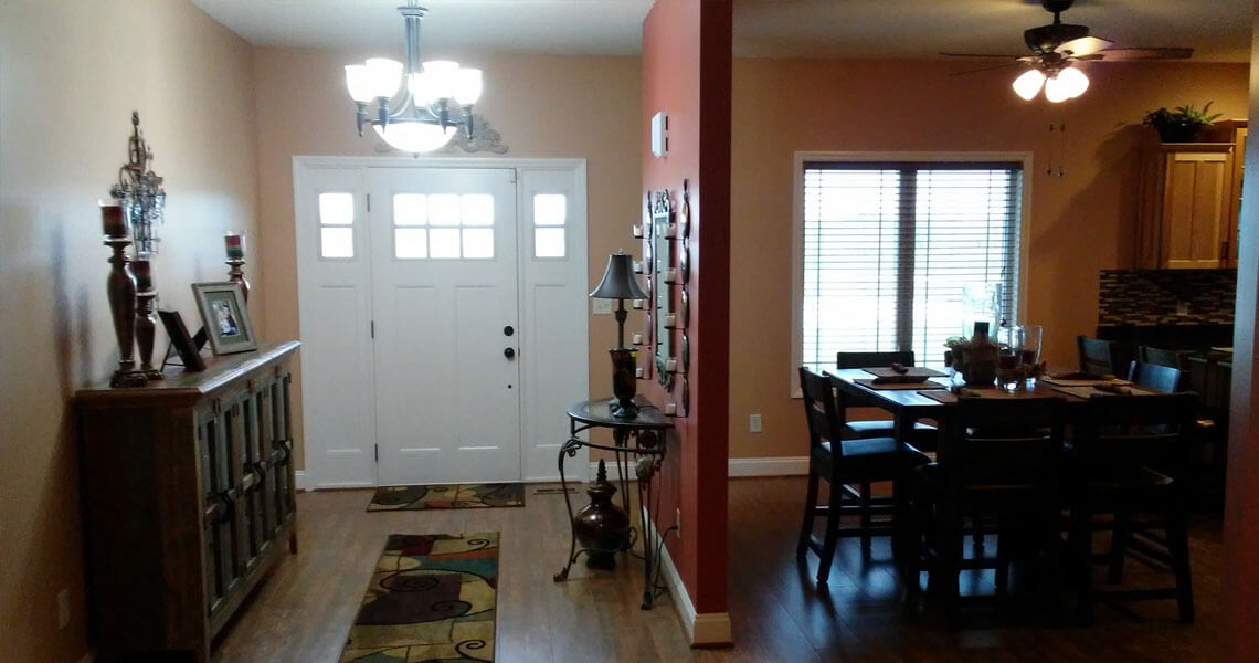 A cozy entryway and dining area with warm lighting. The entry has a white door and a decorative console, while the adjacent dining room features a table set for six under a ceiling fan, near a window with blinds.
