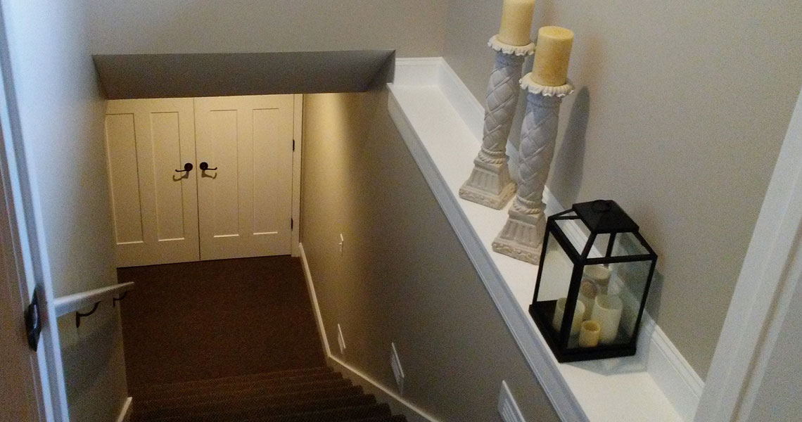 A carpeted staircase leads down to a set of double doors. On the right side, a shelf holds two tall candles on decorative stands and a black lantern with candles inside. The walls and trim are painted in neutral tones.