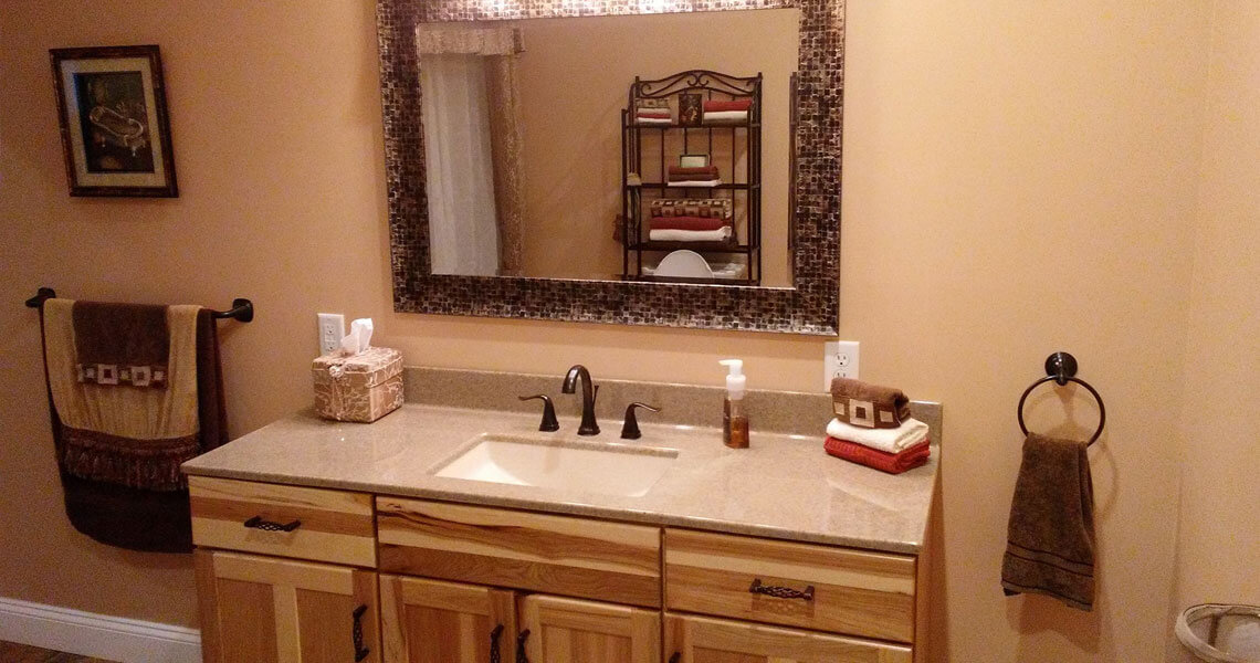 A bathroom with beige walls features a large mirror with a mosaic frame above a wooden vanity with a granite countertop. The sink has a black faucet, and there are towels and toiletries neatly arranged. A towel hangs on a nearby rack.